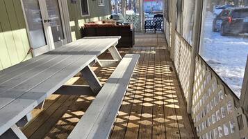 A covered outdoor seating area with picnic tables and a chair, a wooden deck, and a view of a red building through the windows.