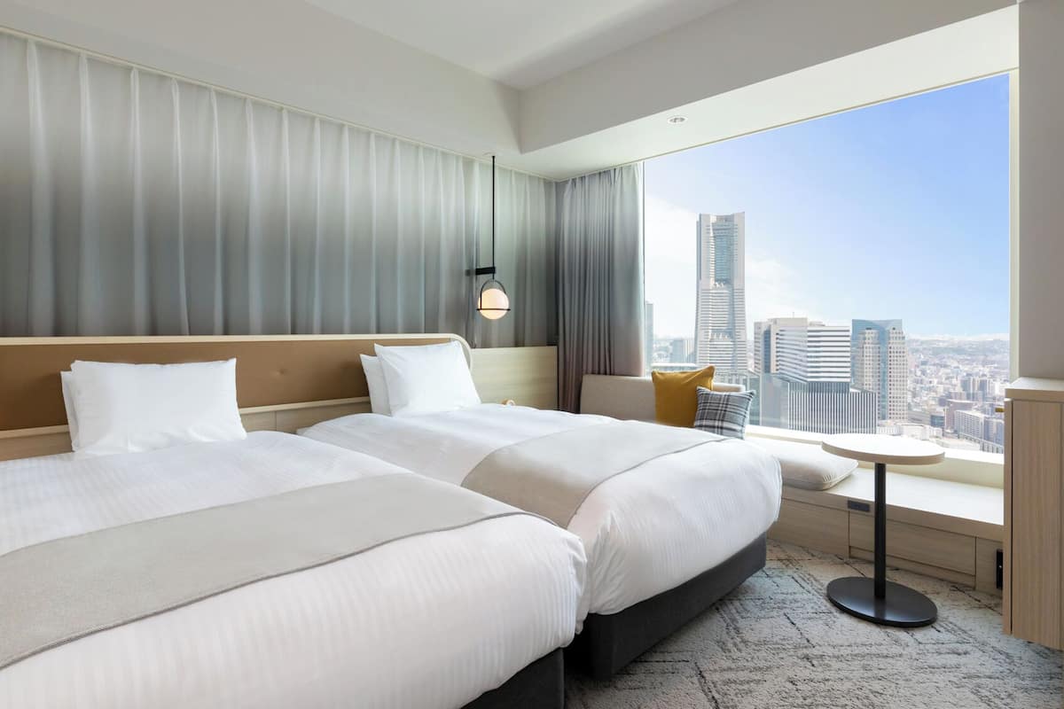 Une chambre d’hôtel avec deux lits, une petite table et une vue sur la ville, avec le panorama urbain visible par la fenêtre.