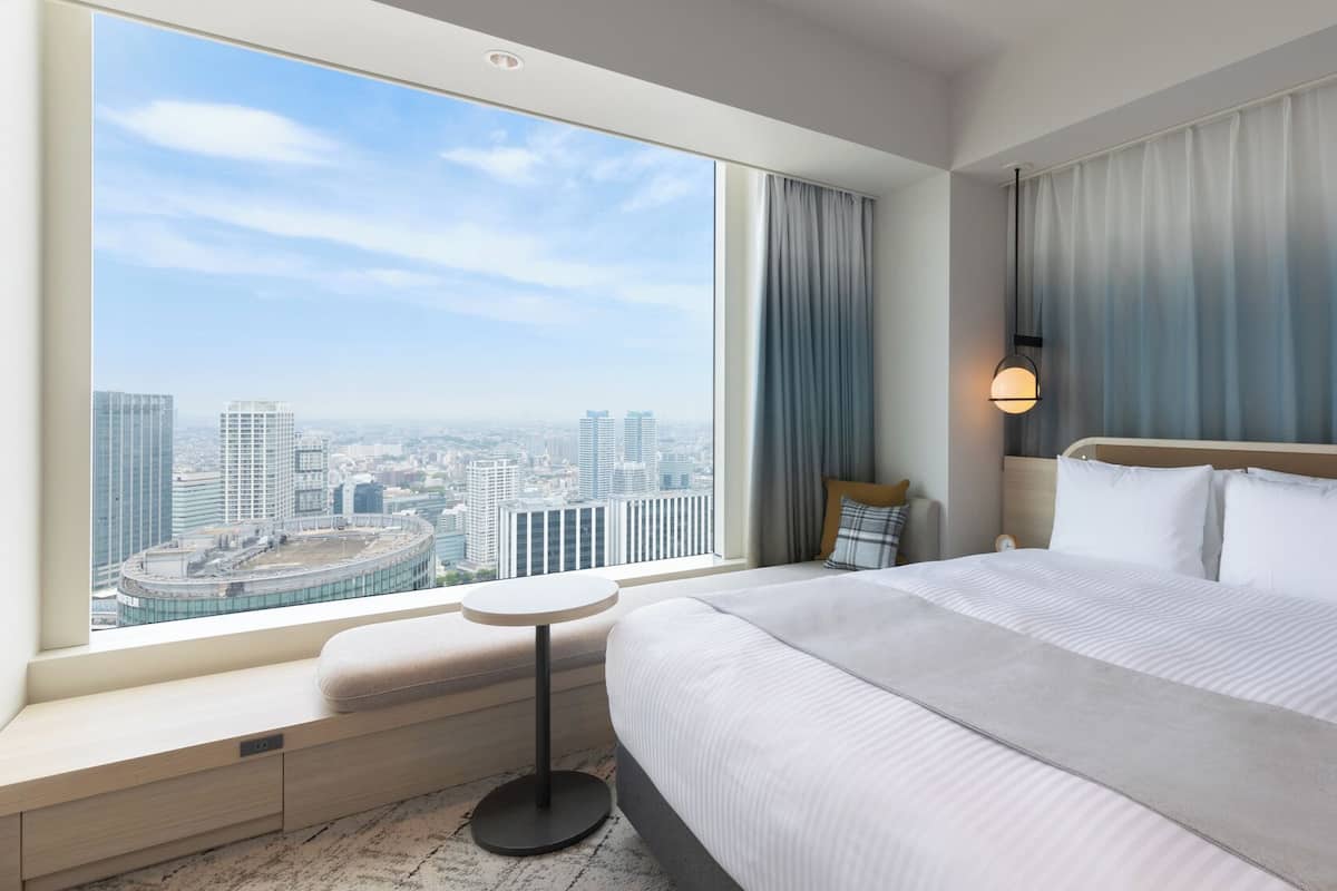 Une chambre d’hôtel avec un grand lit, une table de chevet, une petite table ronde, une commode, une lampe et une vue sur le paysage urbain à travers de grandes fenêtres.