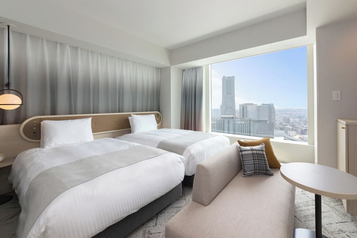 Une chambre d’hôtel avec deux lits, un bureau, une chaise, une télévision et une grande fenêtre donnant sur la ville.