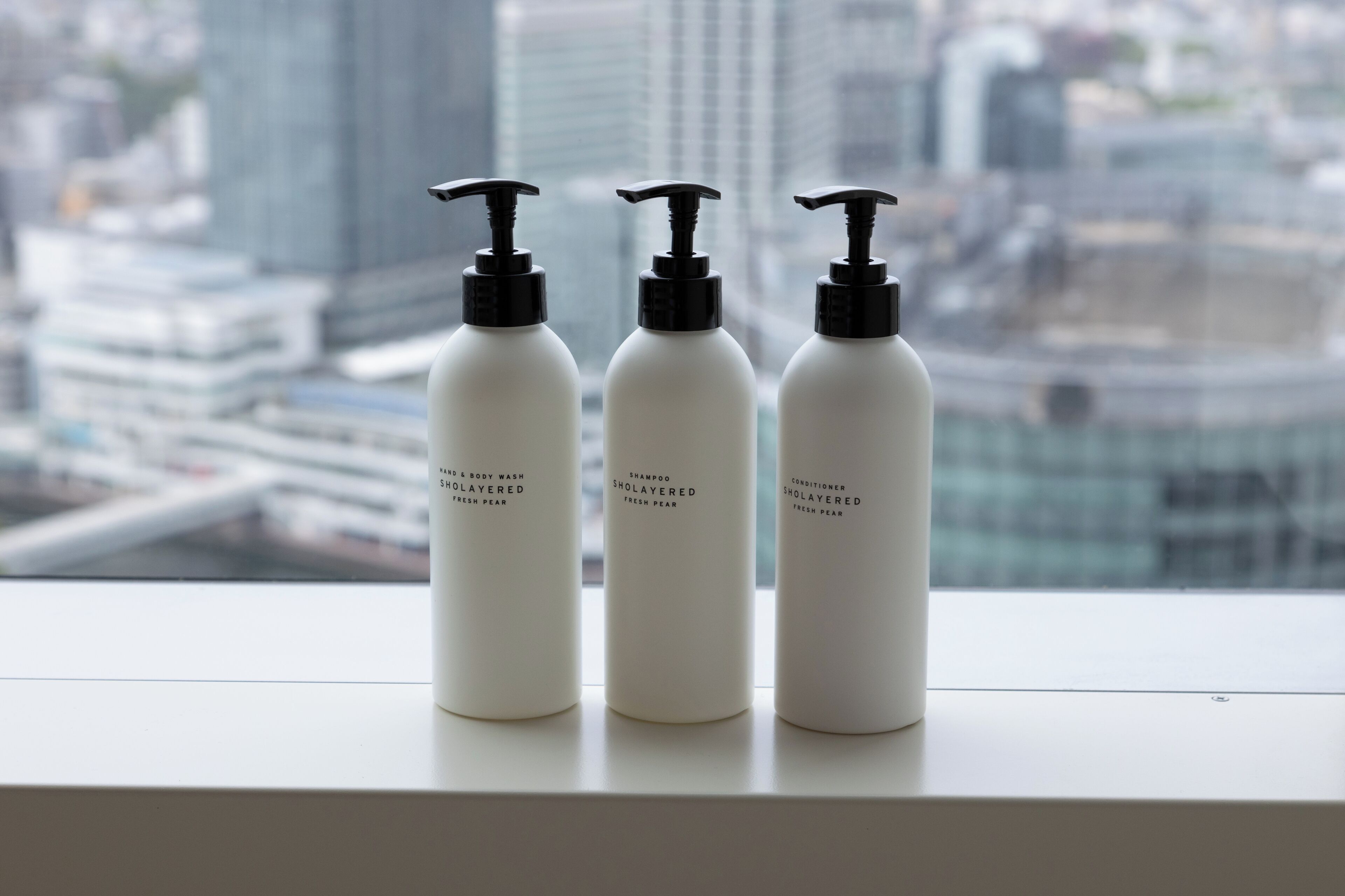 Three white bottles with black pumps on a windowsill, labeled "SHOALAYERED FRESH PEAR," with a cityscape view in the background.