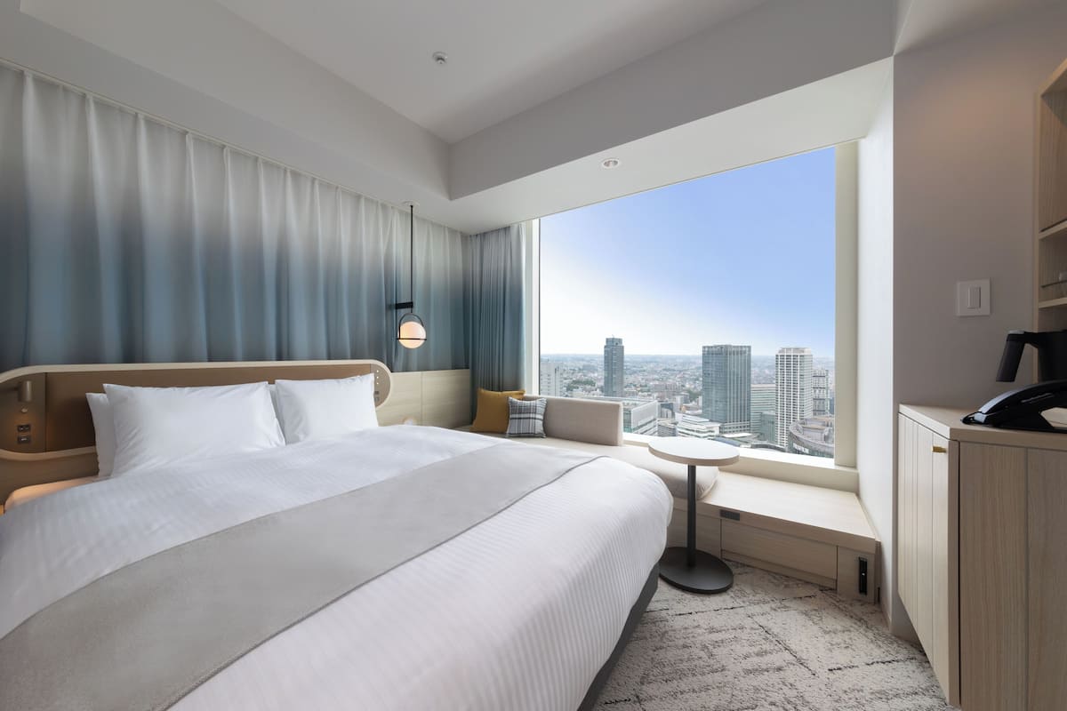 Une chambre d’hôtel moderne dotée d’un grand lit, d’un petit coin salon, d’un bureau et d’une grande fenêtre donnant sur la ville.