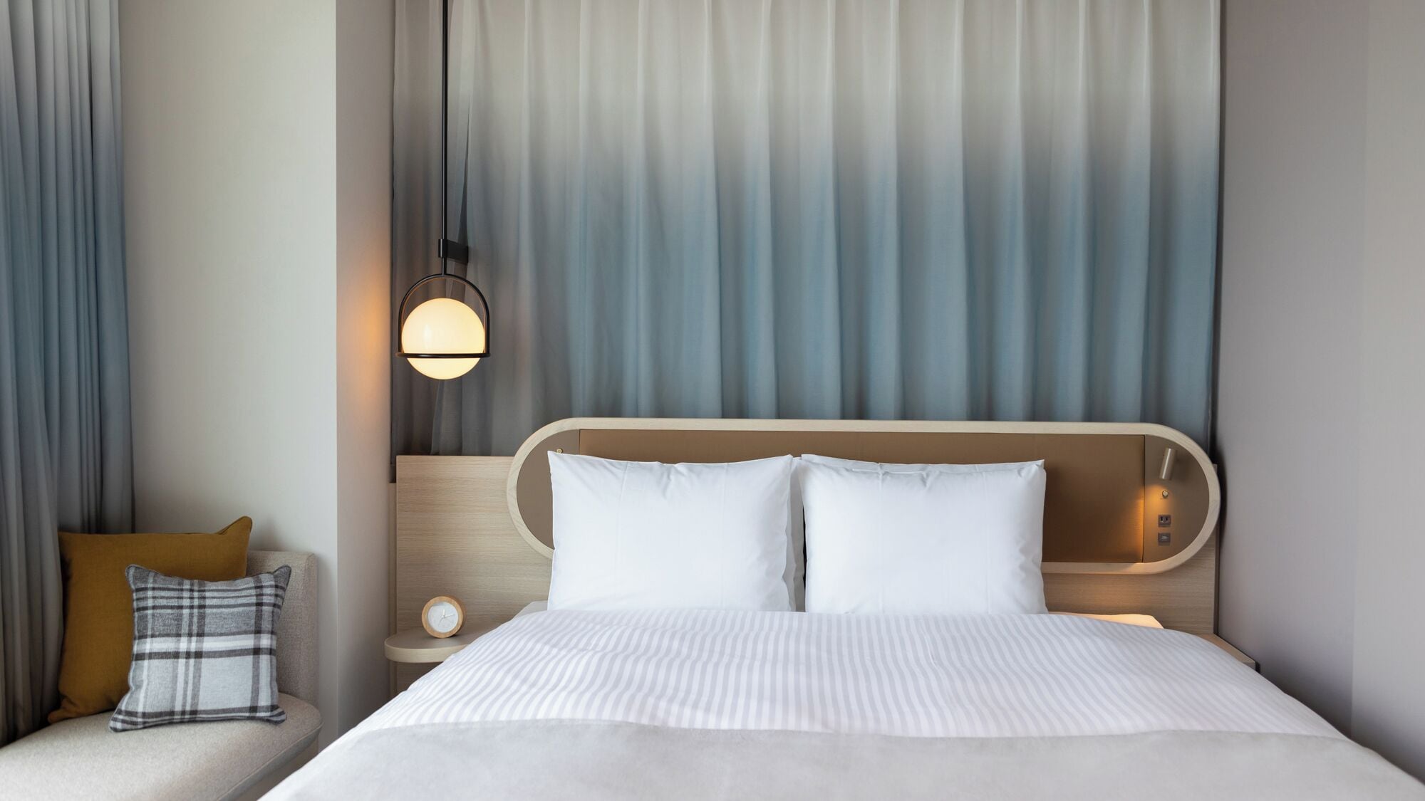 Habitación de hotel con cama, mesita de noche, lámpara y vistas al exterior a través de cortinas.