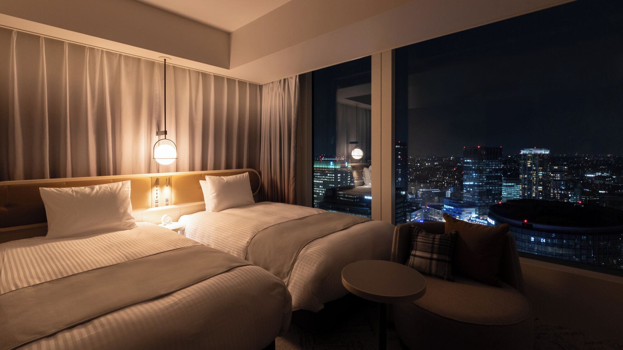 Habitación de hotel con dos camas, una mesita de noche, una silla y un ventanal con vistas a la ciudad.