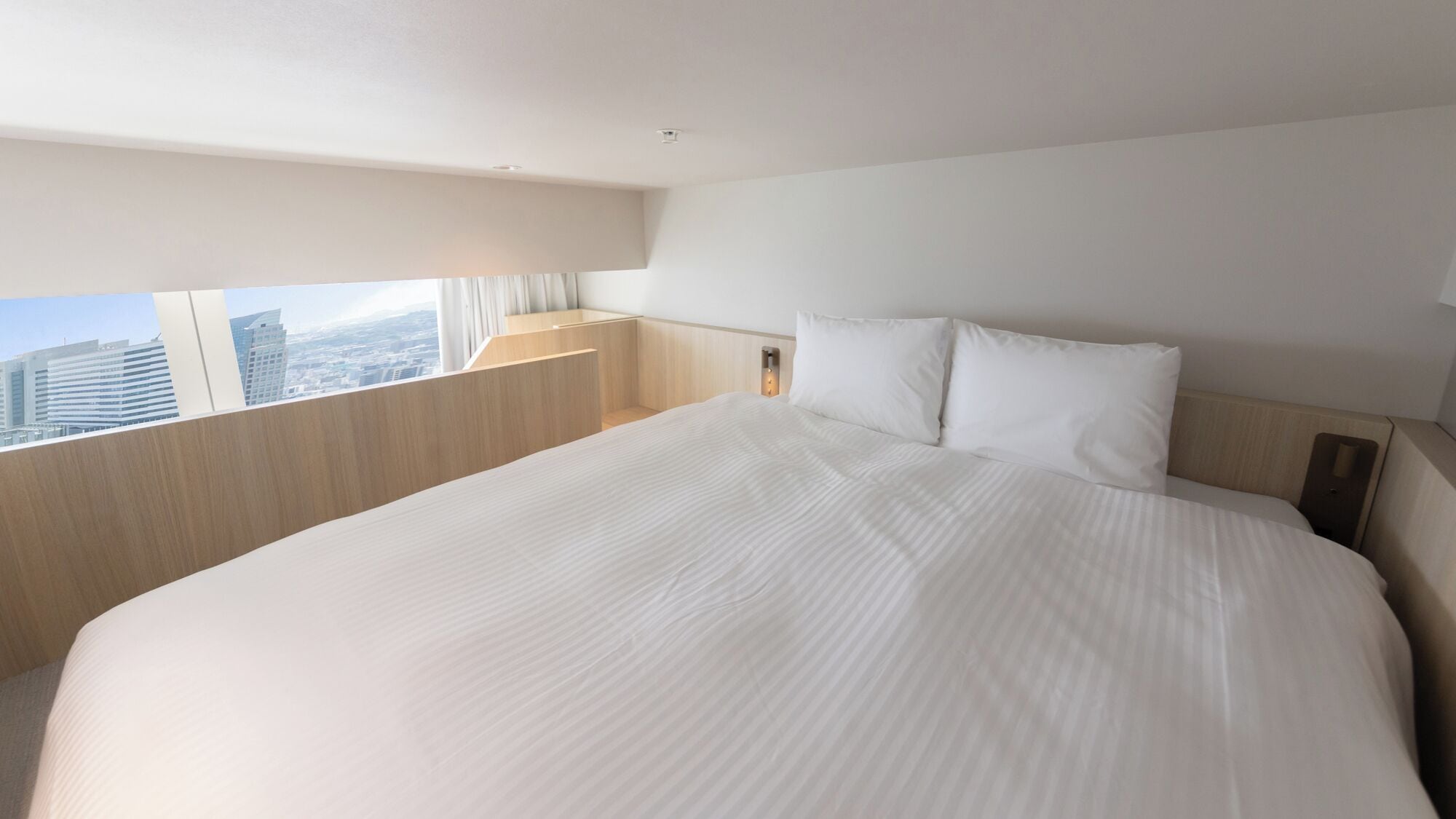 A hotel room with a large bed, a nightstand, a lamp, and a window with a city view.