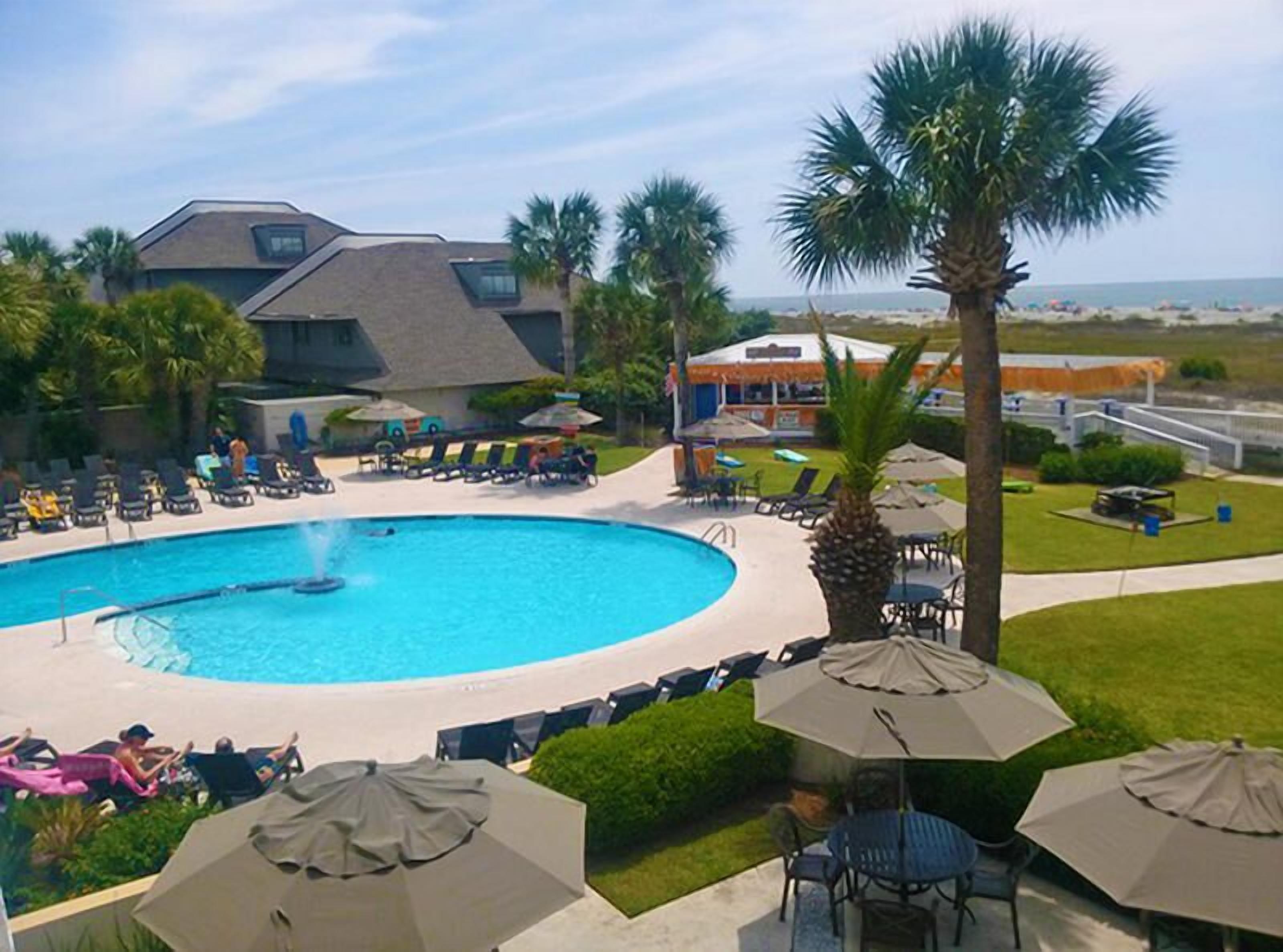A pool area with lounge chairs and umbrellas, surrounded by palm trees and a building with a sloped roof.