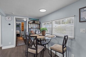 A dining area with a round table, chairs, a clock, a bookshelf, and a window with blinds.