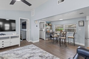 A modern living room with a flat-screen TV, a dining area with a table and chairs, and a kitchen area with a stove and cabinets.