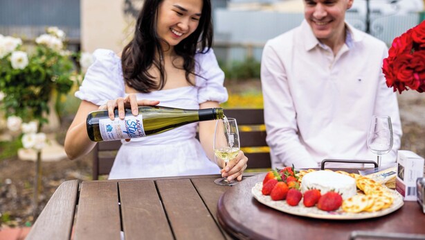Een eettafel met een fles wijn die in een glas wordt gegoten, een bord met aardbeien en kaas, en een boeket rode rozen.