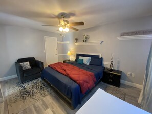 A bedroom with a bed, a chair, a nightstand, a ceiling fan, a lamp, and a sign on the wall.