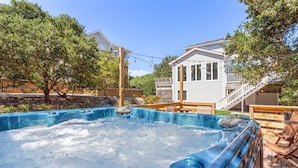 A hot tub with a wooden deck and chairs, surrounded by trees and a house with a balcony. - Mighty Duck: Hot Tub, Fire Pit, Game Room, Duck OBX (Duck)