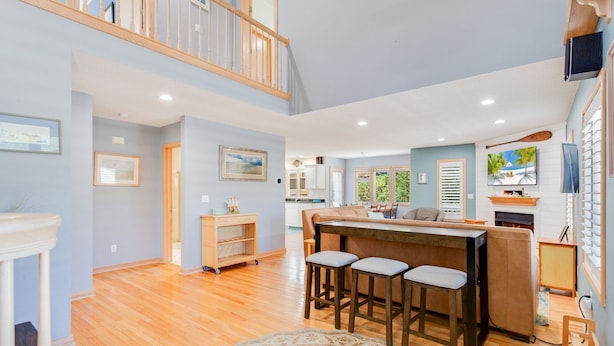 A spacious living area with a wooden floor, a dining table with bar stools, a kitchen in the background, and a fireplace. - Mighty Duck: Hot Tub, Fire Pit, Game Room, Duck OBX (Duck)