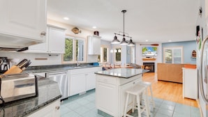 A modern kitchen with white cabinetry, a granite countertop, and a central island with stools. There is a dining area with a wooden table and chairs, and a living room with a sofa and a fireplace. - Mighty Duck: Hot Tub, Fire Pit, Game Room, Duck OBX (Duck)