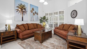 Ein Wohnzimmer mit zwei braunen Sofas, einem Holztisch und einem gemusterten Teppich. Zwei Lampen stehen auf Beistelltischen, und ein großes Fenster mit Jalousien.