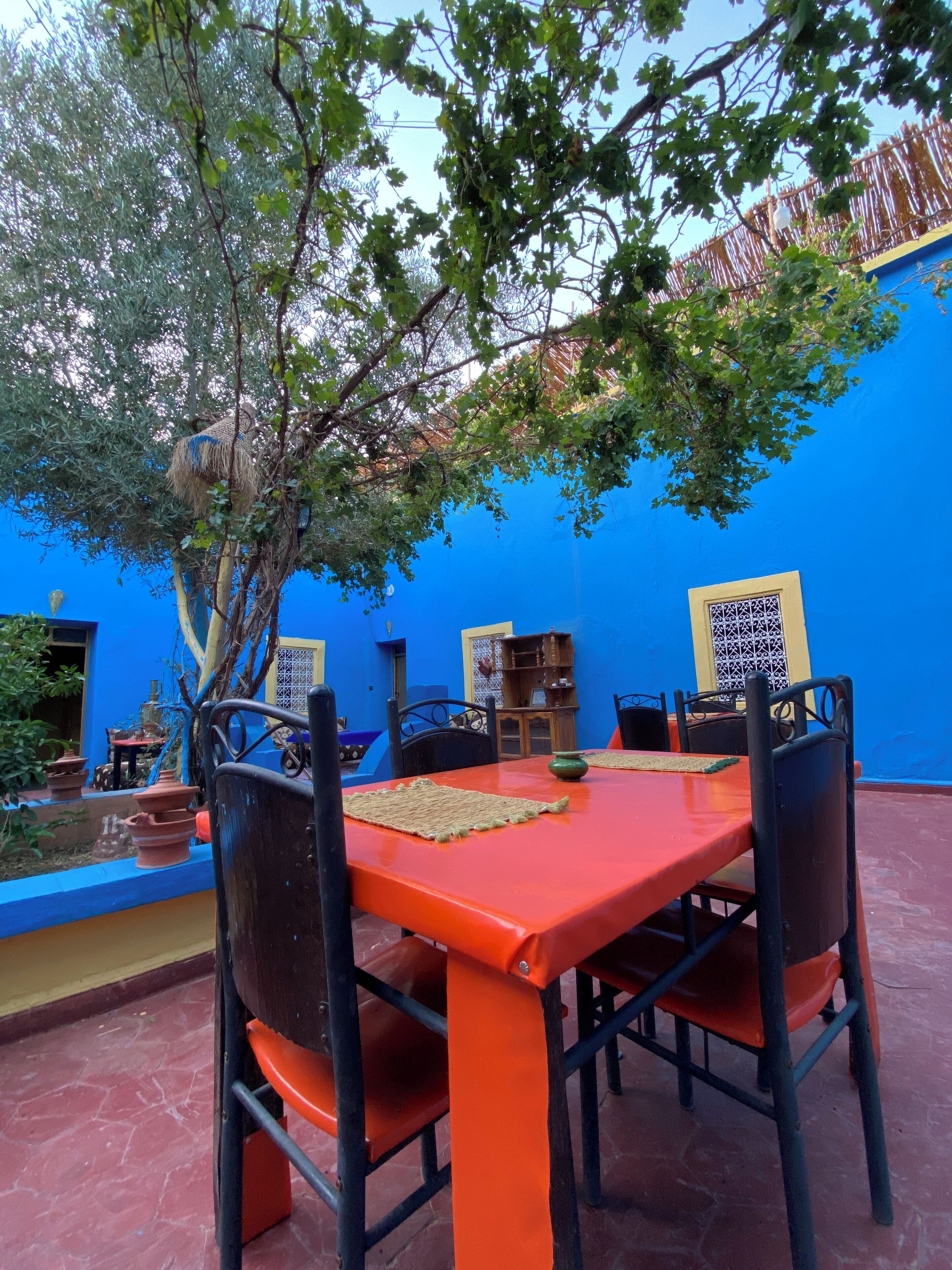 An outdoor dining area with a red table and black chairs, surrounded by a blue wall and a tree.