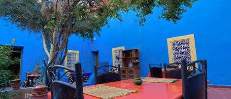 An outdoor dining area with a red table and black chairs, surrounded by a blue wall and a tree.