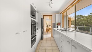 A modern kitchen with white cabinets, stainless steel appliances, and a large window overlooking a balcony. - White Sands House by Experience Jervis Bay (Vincentia)