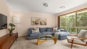 A modern living room with a sectional sofa, a glass coffee table, a wooden sideboard, and a large window offering a view of trees. - White Sands House by Experience Jervis Bay (Vincentia)