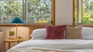 A bedroom with a bed, pillows, a lamp, a small table with a plant, and a window with a view of trees. - White Sands House by Experience Jervis Bay (Vincentia)