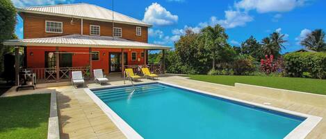 A pool with a wooden deck, a red building with a metal roof, and a clear blue sky.