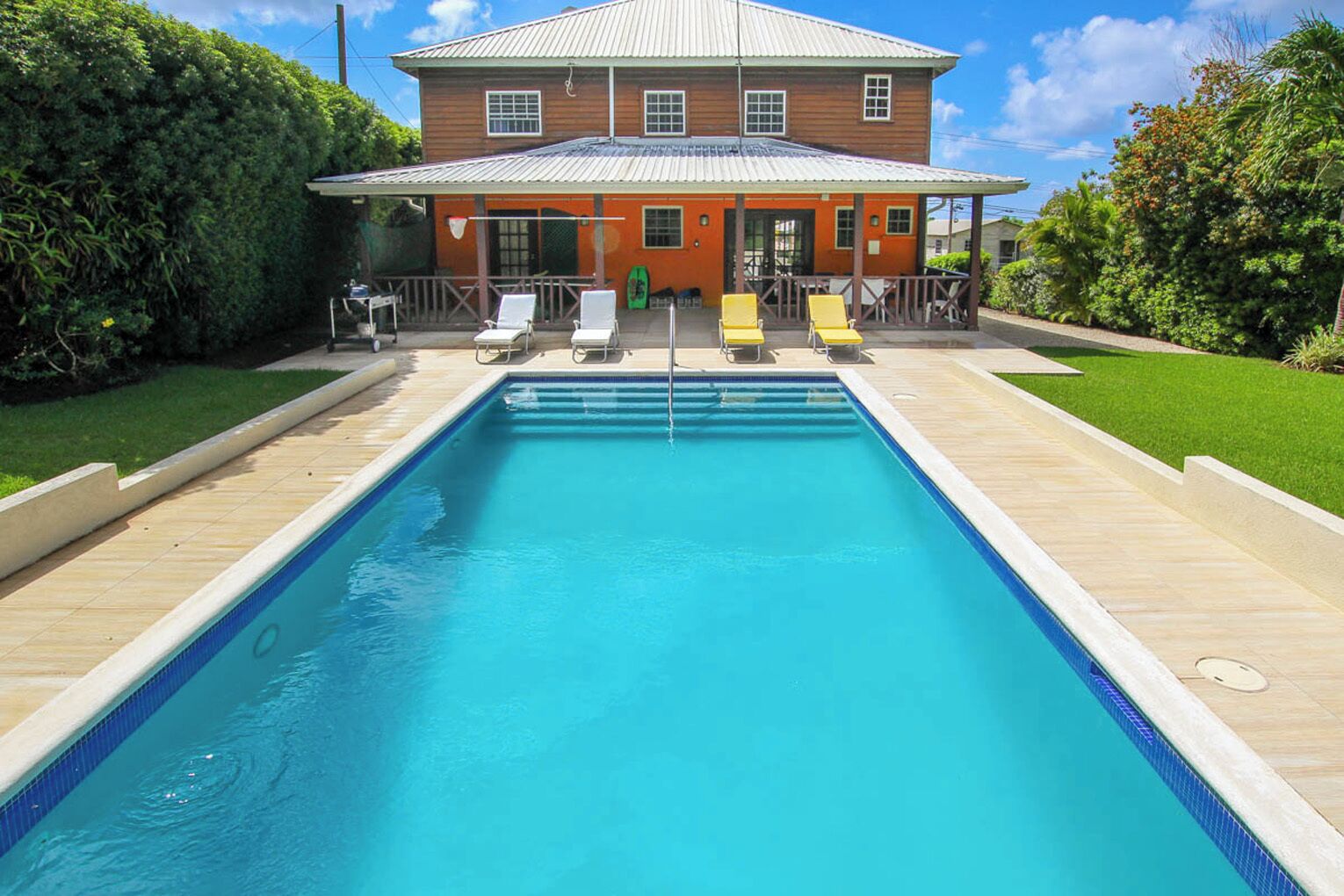 Una piscina rettangolare con un bordo in legno, una casa sullo sfondo e cielo sereno.