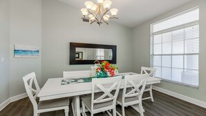 A dining room with a white table and chairs, a chandelier, a large window with blinds, and a painting on the wall. - Enjoy this lovely updated 4-bedroom, 2.5-bath waterfront home 2.5 miles from Tigertail Beach (Marco Island)