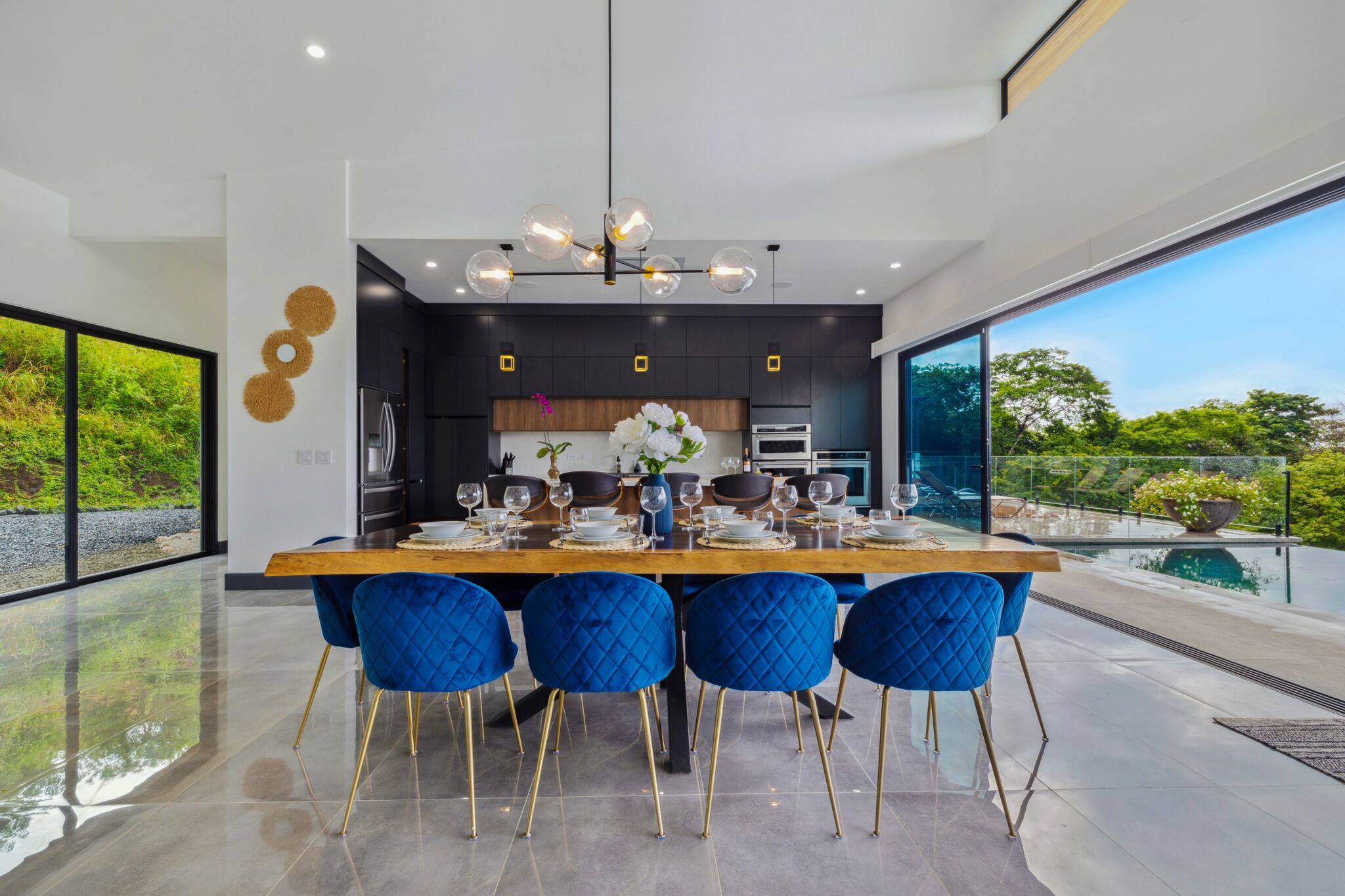 Cocina moderna con una isla grande, sillas azules y un área de comedor con vistas al exterior.