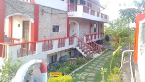 Un edificio de dos pisos con fachada roja y blanca, un balcón en el nivel superior y una pequeña zona ajardinada con macetas.