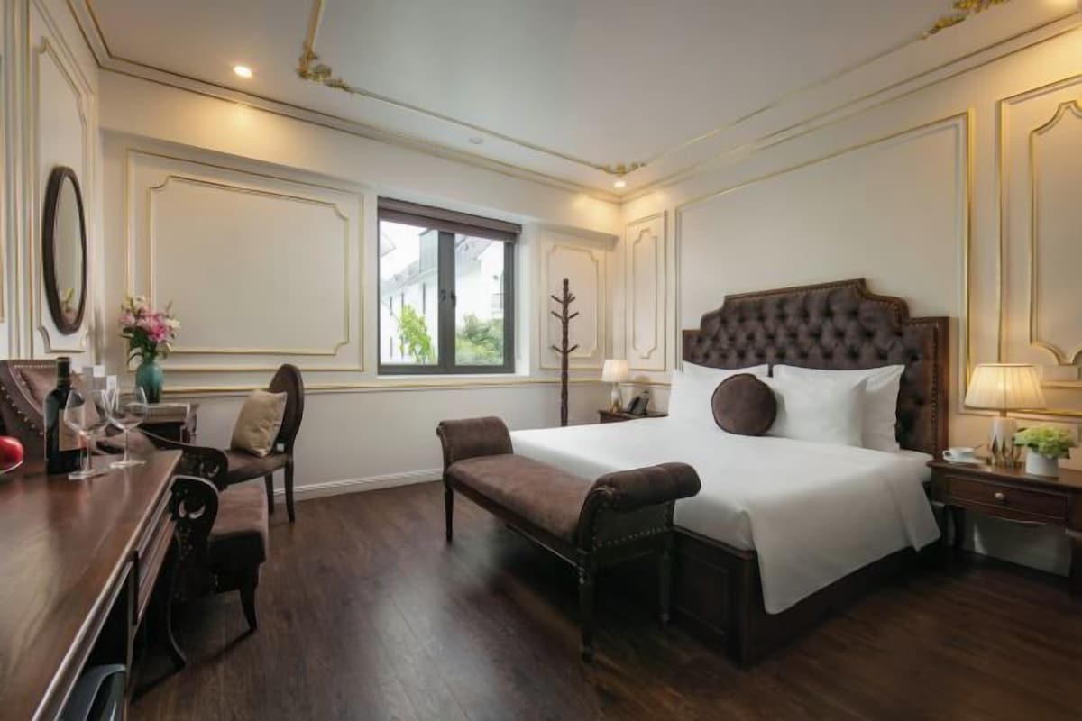 A hotel room with a large bed, a desk with a chair, a window, and a nightstand with a lamp.