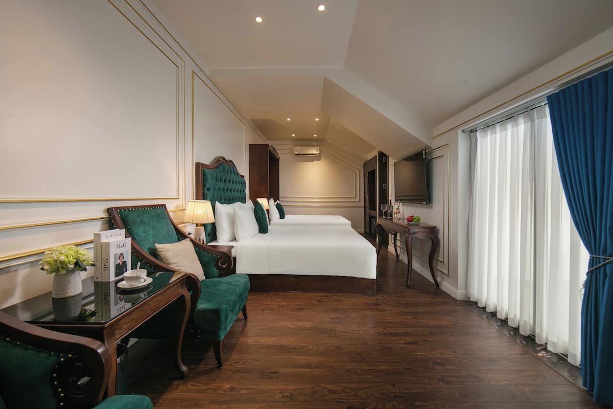 Een hotelkamer met een groot bed, een zithoek met een groene fauteuil, een kleine tafel met een vaas en boeken, en een televisie.