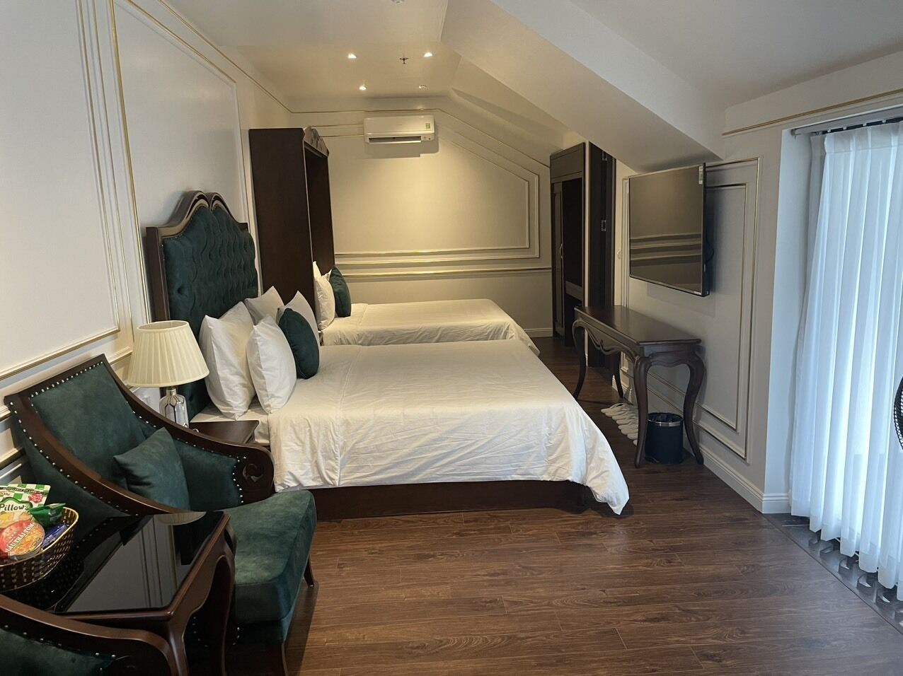 A hotel room with two beds, a sitting area with a chair and a table, a TV, and a window with curtains.