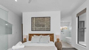 Un dormitorio moderno con una cama de madera, un ventanal grande y un baño a la vista en el fondo.