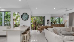 Una cocina moderna con una isla central, taburetes de bar, un comedor con mesa y sillas de madera, un sofá blanco y un reloj de pared grande.