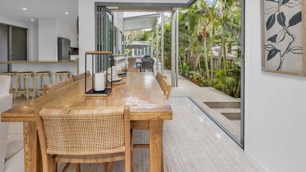 Un comedor moderno con mesa de madera, sillas de mimbre, una pared divisoria de vidrio y vistas a un jardín tropical.