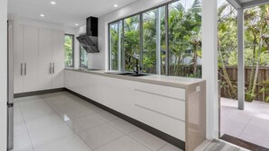 Una cocina moderna con armarios blancos, una campana extractora negra y un amplio ventanal con vistas a un jardín tropical.