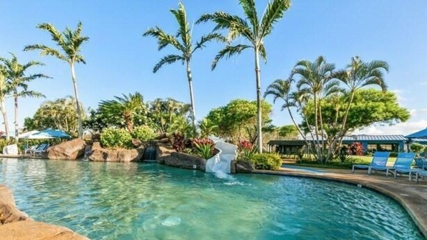 Ein klares Schwimmbecken mit Wasserfall, umgeben von Palmen und Liegestühlen.
