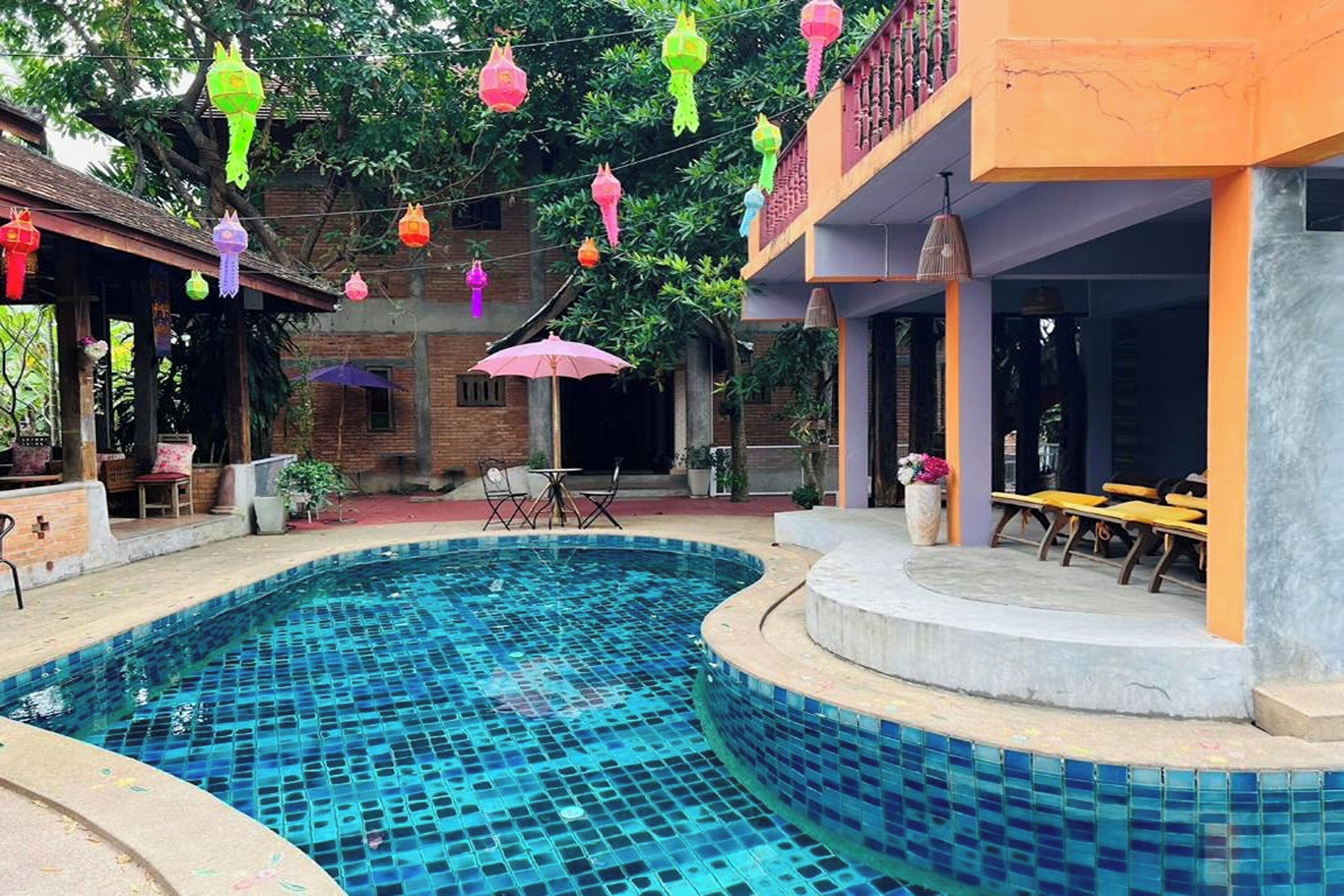 A swimming pool with tiled flooring, surrounded by a covered seating area with chairs and tables, and a building with a balcony.