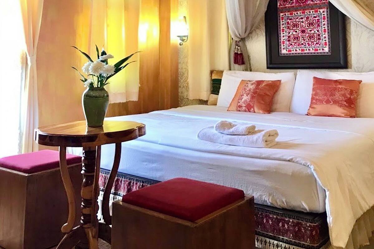 A hotel room with a bed, a small table with a vase of flowers, and a red ottoman.