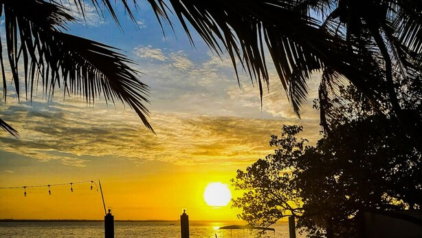 Une vue sur le coucher de soleil au-dessus d’une étendue d’eau calme, avec un bateau amarré près du rivage, entouré de palmiers et sous un ciel dégagé.
