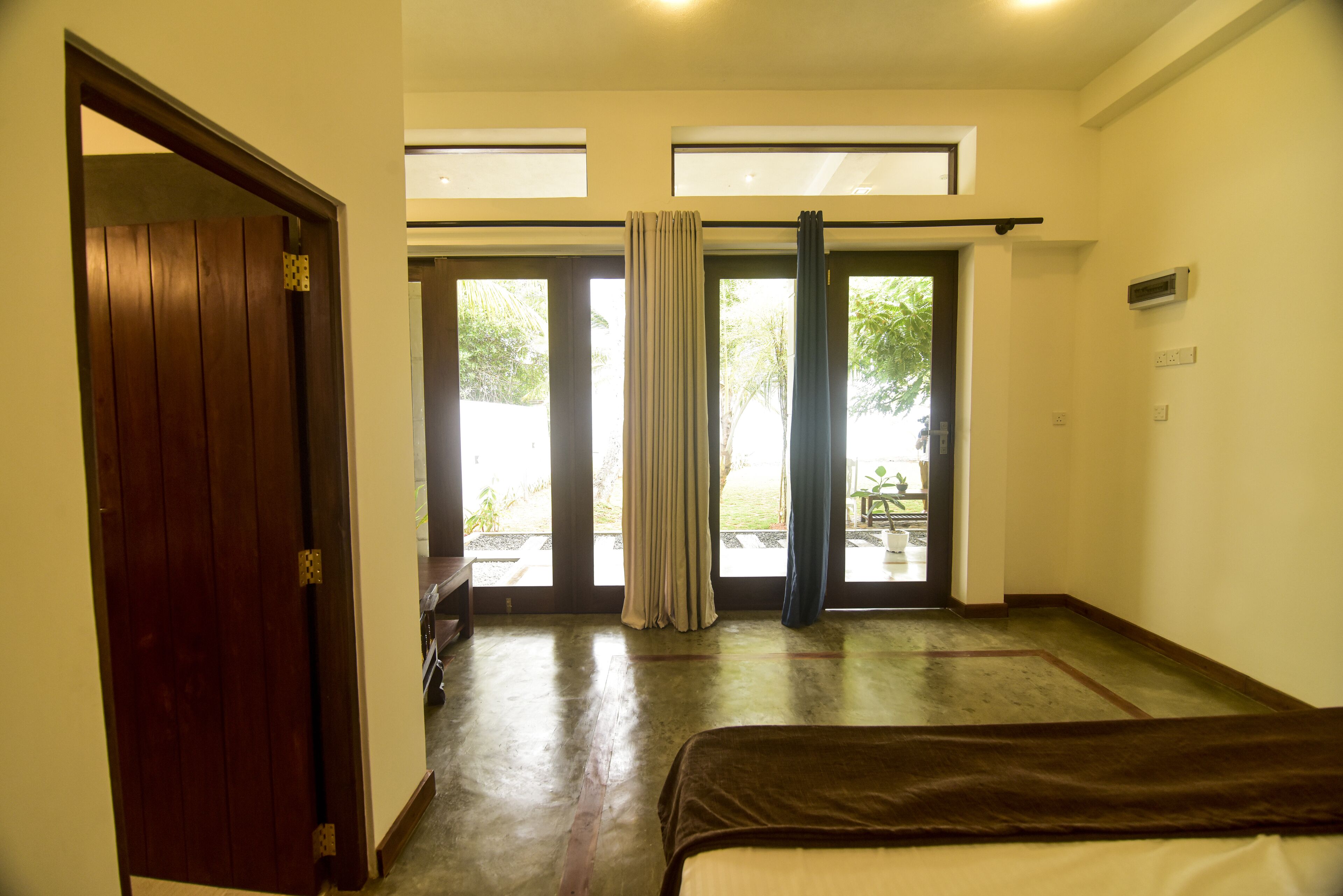 A room with a bed, a wooden door, and a view of the outside through large windows.