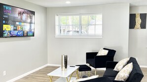 A modern living room with a flat-screen TV, black sofas, a white coffee table, and a window with blinds. - Charming 3BR/2BA Near Galleria/NRG, Top Hospitals & LA Fitness (Houston)