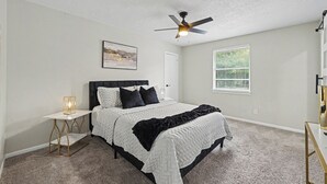 Une chambre à coucher comprenant un lit, une table de chevet, un ventilateur de plafond, une fenêtre avec des stores et une porte.