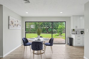 Une cuisine moderne avec des meubles blancs, un coin repas comprenant une table ronde et quatre chaises, et une grande porte-fenêtre coulissante donnant sur un jardin.