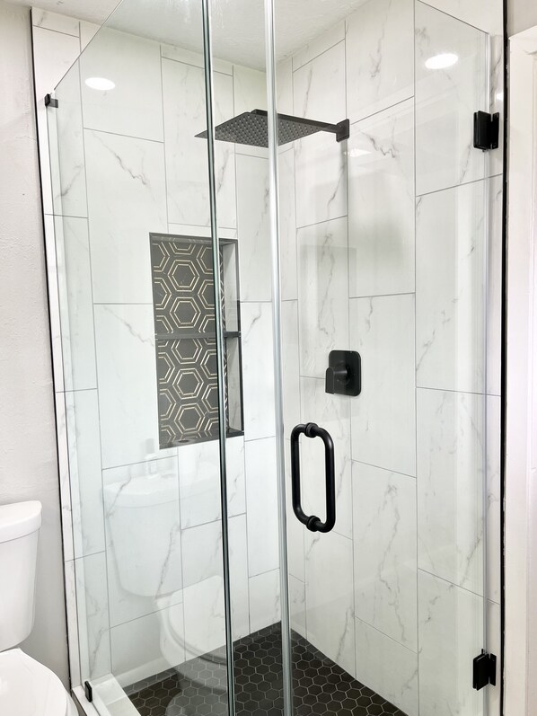 Un baño moderno con ducha de vidrio, azulejos de mármol blanco y un revestimiento de pared con diseño geométrico.