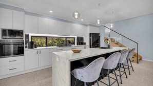 A modern kitchen with white cabinetry, a large island, bar stools, and a view of greenery through a window. - Heather - Pet Friendly Merimbula (Merimbula)