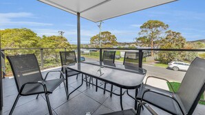 A balcony with a glass table and chairs, offering a view of a street with cars and trees. - Heather - Pet Friendly Merimbula (Merimbula)