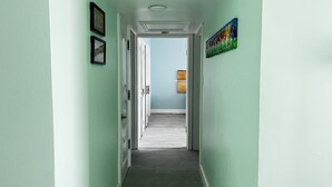A hallway with light green walls, a wooden floor, and a door leading to another room. There are framed pictures on the wall and a colorful abstract painting. - SEAFLECTIONS - Relax, Reflect & Reminisce on the MUSIC, ART and CULTURE.  (Kingston)