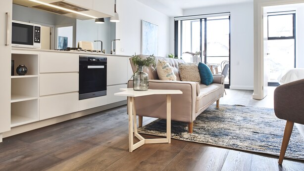 A modern kitchen with built-in appliances, a dining area with a sofa and a coffee table, and a large window providing natural light. - Mega Style Apartments - The Marc (St Kilda)