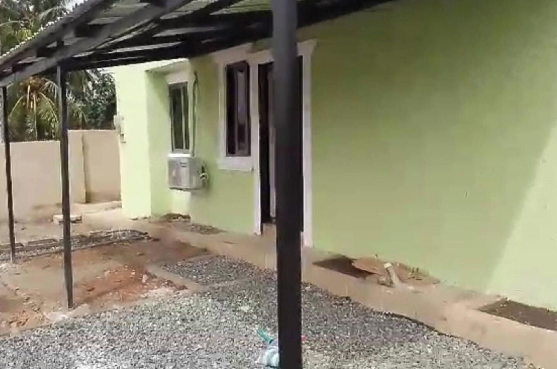 Um edifício com fachada verde, uma janela e uma porta. Há um caminho de cascalho e uma estrutura metálica em primeiro plano.
