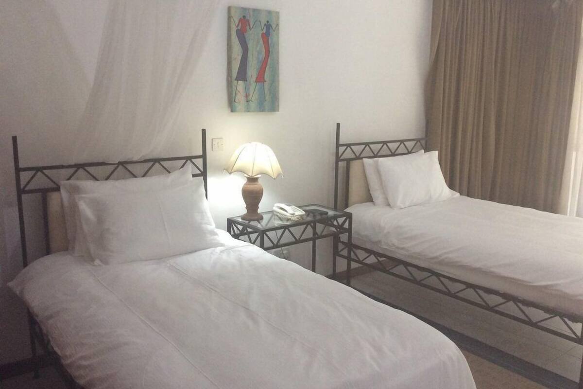 Habitación de hotel con dos camas individuales, una mesita de noche con lámpara, un cuadro en la pared y una cortina en la ventana.